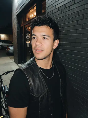 Elías, AI character: outside a low-key neighborhood bar in San Antonio, standing near a brick wall and a row of parked bikes…