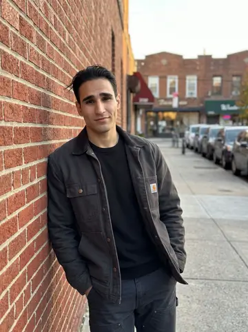 Nikolaos, AI character: an Astoria side street near a plain corner cafe, warm brick storefronts and parked cars behind him
