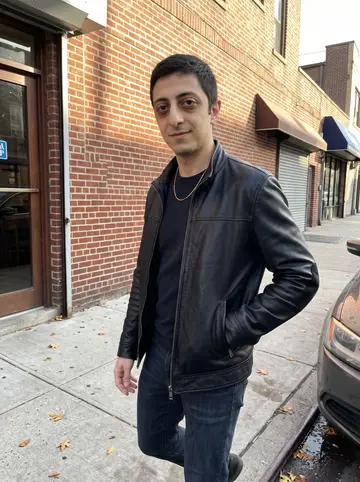 Haris, AI character: a side street in Astoria near a Greek café, brick storefronts and parked cars behind him in late afternoon