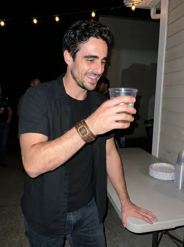 Kimon, AI character: outside the motorcycle club clubhouse during a humid summer-night charity cookout, folding tables and…