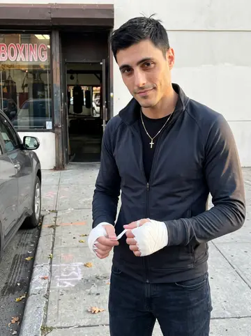 Anastasios, AI character: outside the boxing gym on Ditmars Boulevard in Astoria, just after training, gym windows and parked cars…
