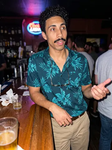 Theo, AI character: a corner bar on Frenchmen Street in the Marigny late on a Friday, a neon beer sign and a crowded wooden…