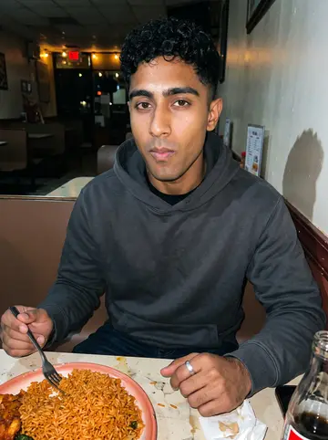 Segun, AI character: a casual Nigerian spot in Houston after closing the shop, sitting at a small table with jollof rice and…