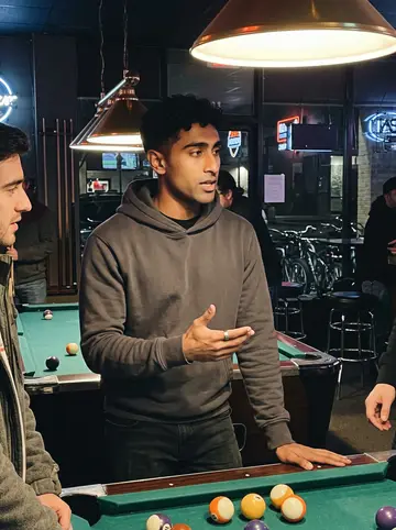 Segun, AI character: a small Houston pool hall with two shop guys half-cropped beside him, bikes visible through the front window