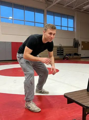 Jesse, AI character: a high-school wrestling room in Oklahoma after practice, red-and-white mats, a folded bleacher and a…