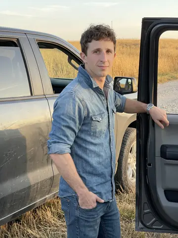 Sawyer, AI character: a gravel pullout beside a Kansas highway in the Flint Hills, tall dry golden grass, leaning on the open…