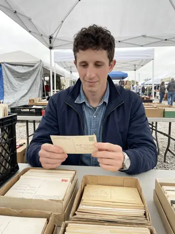 Sawyer, AI character: a flea market stall under a white canopy tent, a folding table covered in boxes of old postcards, in a…