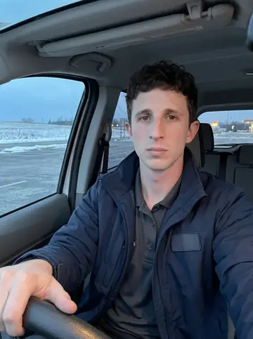 Sawyer, AI character: parked at a highway rest stop in the driver's seat of the company van at dusk, snow-dusted fields through…