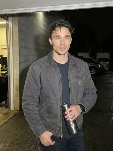 Edwin, AI character: under the tow yard awning at drizzly dusk, wet pavement and parked trucks fading into the dark behind him