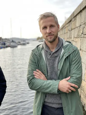 Sindre, AI character: the stone sea wall in Nordnes park on the Bergen waterfront, the grey fjord and moored boats behind him