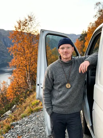 Halvard, AI character: the narrow road above the Hardanger fjord, leaning against the open door of his white work van, orange…
