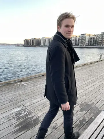 Stig, AI character: the wooden boardwalk at Sørenga on the Oslofjord, calm grey-blue water and apartment blocks behind him