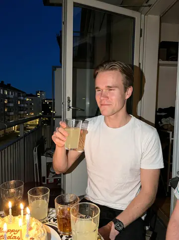 Stig, AI character: the small balcony of his Oslo apartment on a late summer evening, a cake with candles and mismatched…