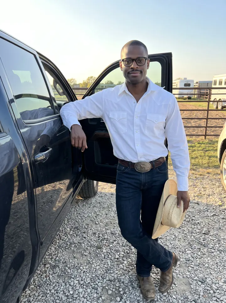 Jamal, AI character: a gravel lot outside a friend's arena in Hempstead, leaning against the open door of his dark pickup after…