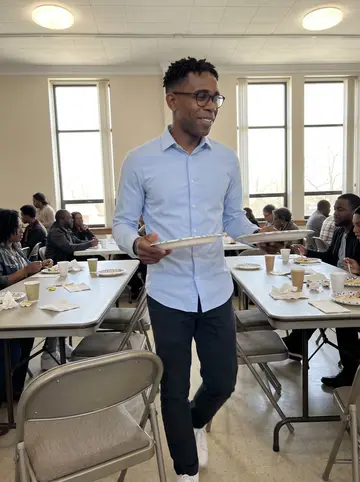 Tyrese, AI character: church fellowship hall in Columbus after Sunday gospel brunch, walking between folding tables with plates…