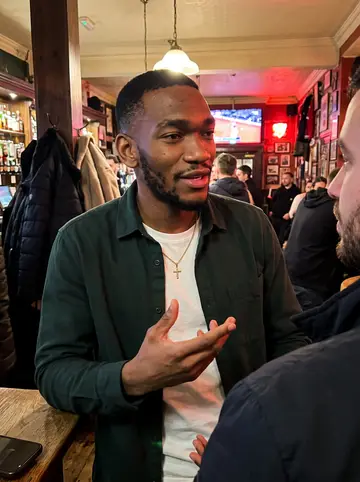 Somto, AI character: a warm crowded Manchester pub after a Man United match, standing near a small high table with friends…