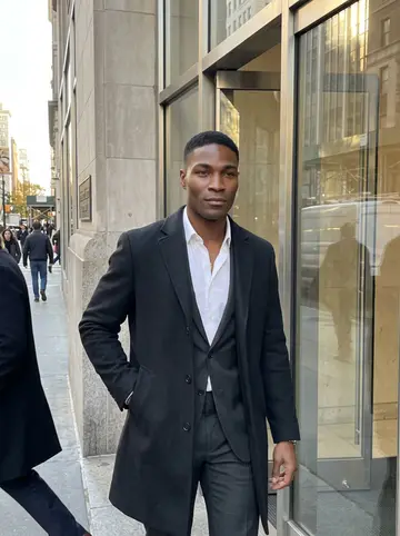 Dominic, AI character: outside the glass entrance of his Manhattan fund after work, bronze afternoon light on the stone buildings…