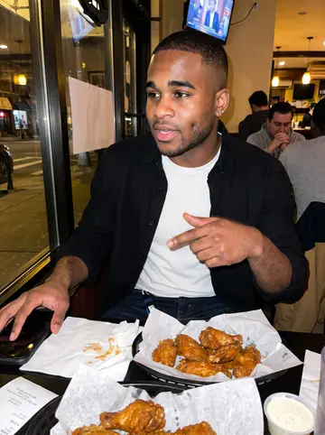 Kendrick, AI character: a crowded neighborhood wing spot he is ranking in his spreadsheet, small table near the front window with…