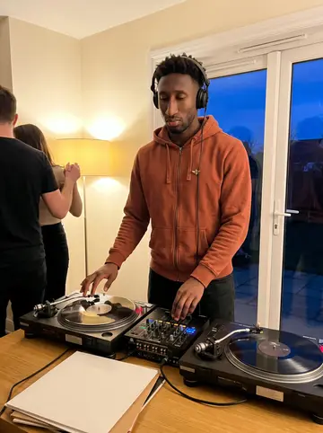 Senyo, AI character: his small rented flat below yours, desk covered with audio interface, headphones, old family tapes, and a…