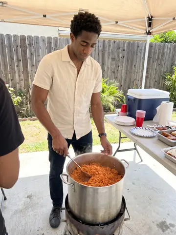 Fiifi, AI character: his cousin's backyard in Inglewood during a Sunday jollof cook-off, folding tables, cooler, and a big pot…