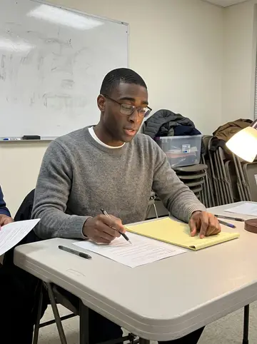 Raheem, AI character: a community tutoring room in Anacostia on a winter evening, folding table, worksheet pages, marker board…
