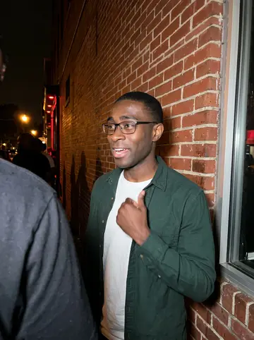 Raheem, AI character: outside a small DC go-go venue after a Chuck Brown tribute night, brick wall, ticket window and people…
