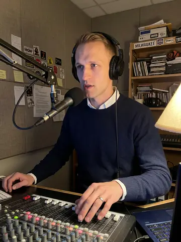 Torben, AI character: inside a small community radio booth in Milwaukee late on Thursday night, mixer board and microphone arm…