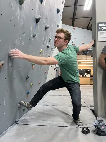 Kilian, AI character: the bouldering gym near his Denver building on a Wednesday night, colored holds on a matte grey climbing wall