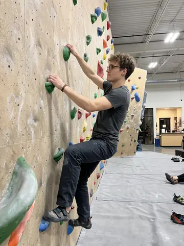 Flavio, AI character: an indoor bouldering gym on a Thursday night, colored climbing holds and padded flooring around him