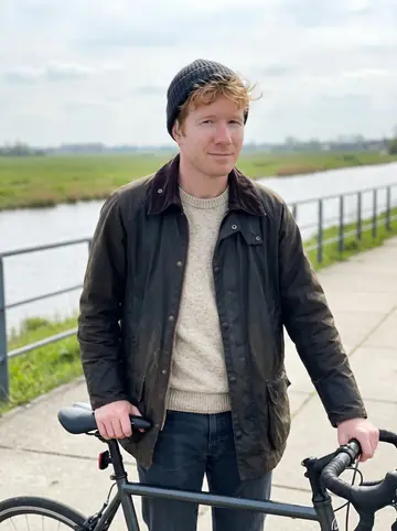 Maarten, AI character: a dike path outside Rotterdam in April, his road bike stopped beside the canal with low green fields…