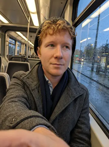 Maarten, AI character: inside a Rotterdam tram at dusk, rainy window glass behind him with streetlights streaking outside