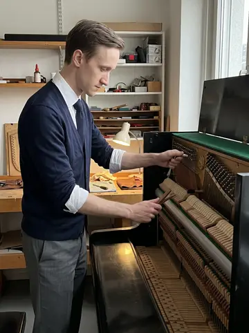 Sandro, AI character: inside the piano workshop in Basel on an overcast autumn afternoon, beside an old upright piano with its…