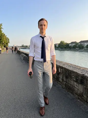 Sandro, AI character: Rhine riverside path in Basel during a warm summer golden hour, low stone wall and river behind him