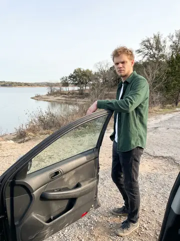 Rens, AI character: parking pullout near a quiet lakeside retreat outside Austin, rental car door open, pale winter lake behind