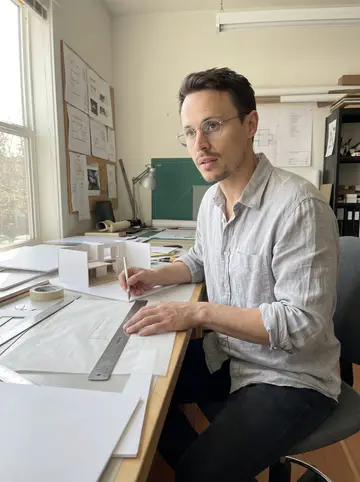Hannes, AI character: his small architecture workroom in Portland, beside a long table covered with tracing paper, foam board,…