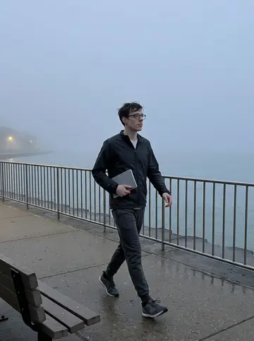 Elia, AI character: the Chicago lakefront path before sunrise, fog over the water and a metal railing along the path