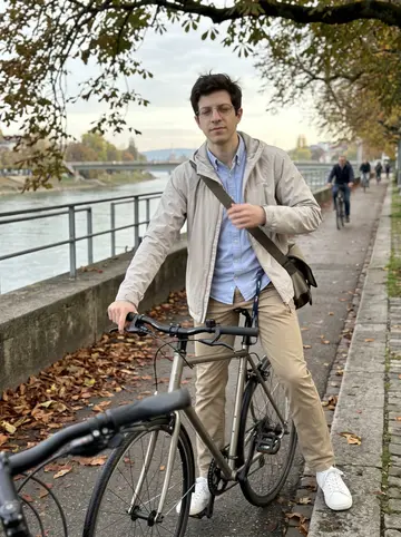 Andrin, AI character: the Rhine cycle path in Basel before work, with damp leaves along the railing and morning commuters behind him