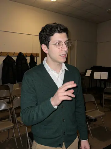 Andrin, AI character: a small community hall after Wednesday yodel choir rehearsal, folding chairs and coat hooks behind him