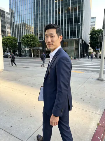 Johnny, AI character: a quiet downtown Los Angeles sidewalk outside a glass office building after work, warm low sun between the…