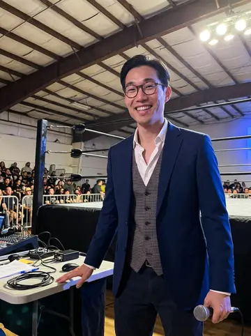 Derek, AI character: inside a small San Jose roller rink during a local wrestling show, standing near the announcer table with…