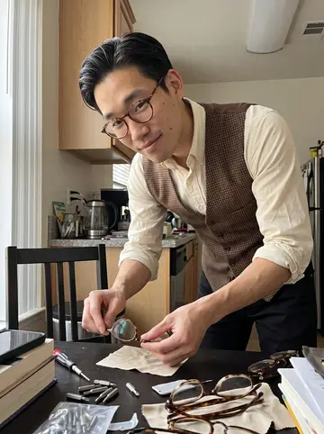 Derek, AI character: his apartment kitchen table in San Jose, covered with small screwdriver bits, lens cloths, and vintage…