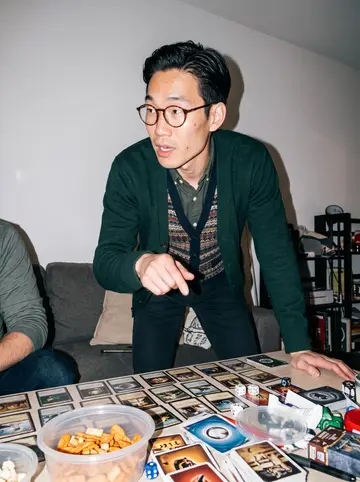 Derek, AI character: a friend's apartment in San Jose during a board game night, crowded table with cards, dice, and snack bowls
