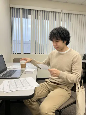 Wilson, AI character: break table in a small Irvine accounting office, laptop, spreadsheet printouts, and a paper coffee cup on…
