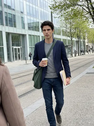 Étienne, AI character: outside a glass office building near Part-Dieu between client meetings, tram tracks and spring trees in…