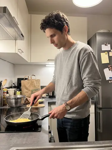 Étienne, AI character: his compact Lyon apartment kitchen at night, pan on the hob, mixing bowl and grocery bag on the counter,…