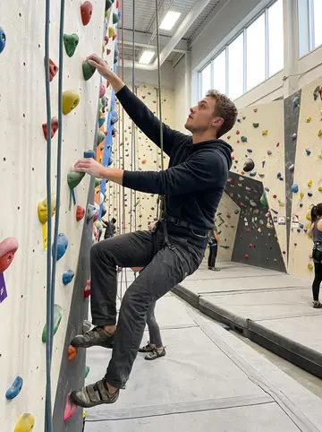 Aymeric, AI character: an indoor rock climbing gym in Montreal on a Wednesday evening, colored holds and rope lanes behind him