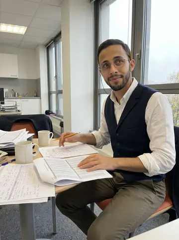 Maxence, AI character: the break area of a Lyon actuarial office, spreadsheet printouts and coffee mugs on a small table