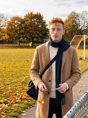 Darragh, AI character: the edge of a community football pitch in Canton, Boston, leaning against a chain-link fence post, fallen…