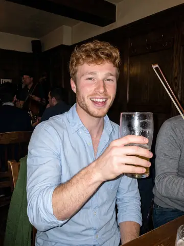 Enda, AI character: a crowded Irish pub in the Annex during a trad session, dark wood wall and blurred musicians behind him
