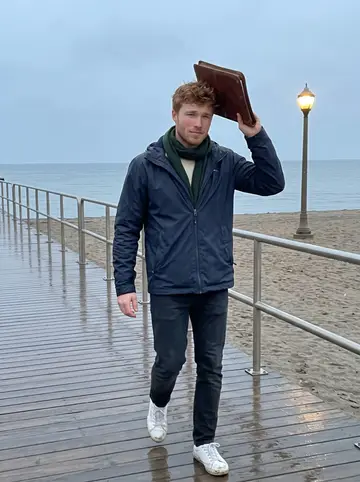 Enda, AI character: the Beaches boardwalk in light drizzle, lake grey-blue behind him and damp sand beyond the railing